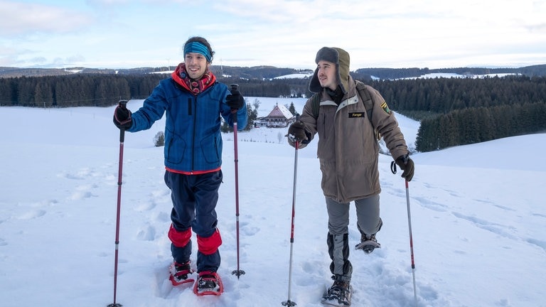Albert und Sebastian wandern mit Schneeschuhen durchs Gelände, im Hintergrund liegt der Fallerhof
