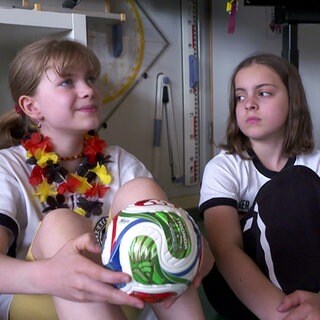 Zwei Grundschülerinnen sitzen auf dem Boden, eine hält einen Fußball in den Händen. Mit einer Broschüre lernen sie mehr über die Fußball-WM.