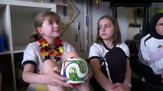 Zwei Grundschülerinnen sitzen auf dem Boden, eine hält einen Fußball in den Händen. Mit einer Broschüre lernen sie mehr über die Fußball-WM.