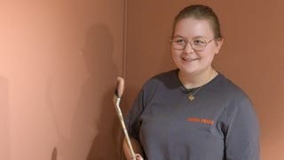 Katharina Bauer steht in einem schwarzen T-Shirt vor eine rötlichen Wand und hält einen hölzernen Pinsel in der Hand. Sie lächelt in die Kamera.