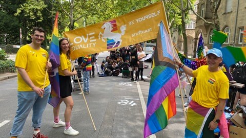 Chiara, Michael und Max sind beim CSD Stuttgart 2025, tragen gelbe Shirts und schauen mit Fahnen in die Kamera.