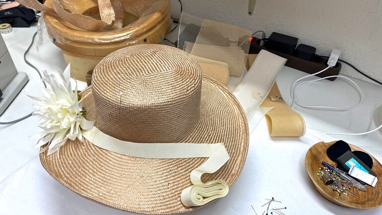 Ein Strohut mit Blumenverzierung auf dem Tisch im Atelier der Modistin - jetzt muss noch das Hutband aufgenäht werden.