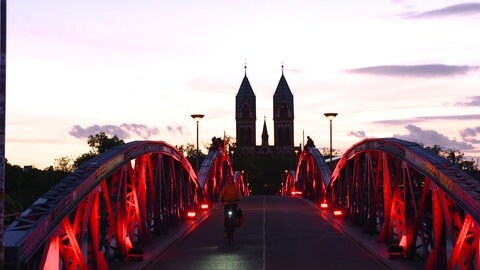 Die Wiwili-Brücke in Freiburg, auch Blaue Brücke genannt, erstrahlt an diesem Abend in rotem Licht. Eine Frau fährt auf einem Fahrrad über die Brücke. 
