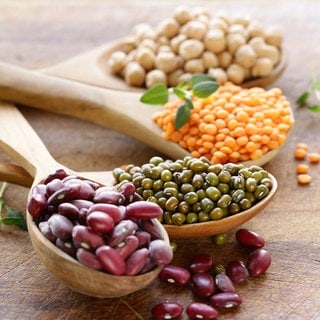 Kichererbsen, rote Linsen, Mungbohnen und Kidneybohnen liegen auf Holzlöffeln auf einer Platte. Wie gesund sind Hülsenfrüchte?