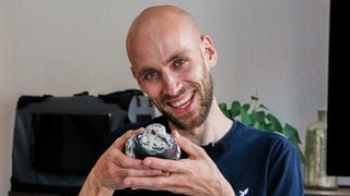 Junger Mann, ohne Haare, mit Bart hält eine Taube in beiden Händen und lächelt in die Kamera.