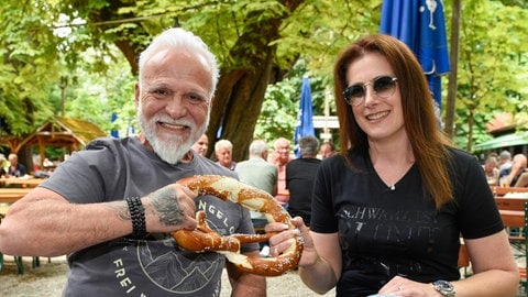 Links sitzt ein Mann mit grauen Haaren und Vollbart in Jeans mit einer Frau mit braunem Haar und Sonnenbrille in einem Biergarten und jeder zieht mit einer Hand an einer riesigen Brezel - Nino de Angelo und Freundin Simone Lux