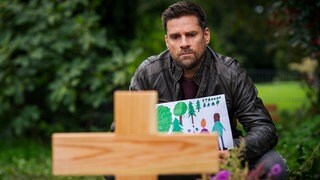 Andreas (Ralph Gassmann) sitzt mit einer Kinderzeichnung in der Hand vor einem Grabkreuz aus HolzKeyvisual