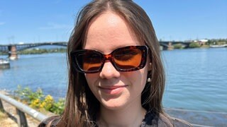 Junge Frau mit Sonnenbrille steht vor Wasser.