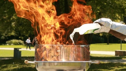 Feuerwehrmann in Schutzanzug schüttet Spiritus auf brennenden Grill mit Stichflamme. Erste Hilfe bei Verbrennungen: Soll man Brandwunden kühlen?