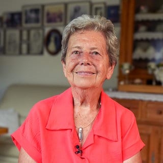 Inge (79) sitzt in ihrem Wohnzimmer und schaut zufrieden nach vorne. Um ihren Hals hängt eine Kette, die sie aus altem Silberbesteck gefertigt hat. Sie hat kurze, graue Haare und trägt eine leuchtend-rosa Bluse.