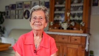 Inge (79) sitzt in ihrem Wohnzimmer und schaut zufrieden nach vorne. Um ihren Hals hängt eine Kette, die sie aus altem Silberbesteck gefertigt hat. Sie hat kurze, graue Haare und trägt eine leuchtend-rosa Bluse.
