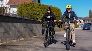 Niklas und Amos fahren jeden Tag mit dem Fahrrad in die Schule. Hier fahren sie gemeinsam durch die Nachbarschaft, hinter ihnen fährt ein Auto.