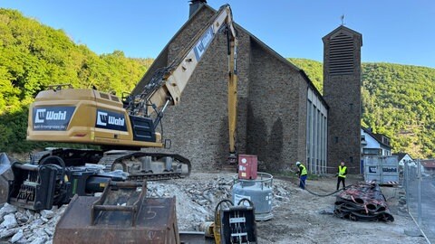 Ein Bagger reißt die St. Andreas Kirche in Ahrbrück ab, ein Bauarbeiter hält einen Wasserschlauch auf die Abrissstelle, um die Staubbildung zu mindern.