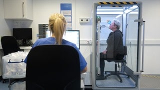 Eine Frau sitzt in einer abgeschlossenen Kabine und atmet durch ein Mundstück. Eine Arzthelferin überprüft an einem Computer die Lungenfunktion der Patientin.