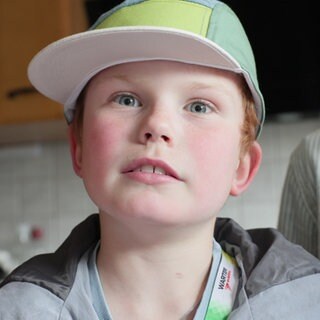 Ben, Spitzname "Lumpi", guckt in die Kamera. Er hat blaue Augen und eine gelb-grüne Kappe, unter der rötliche Haare hervorschauen. Im Hintergrund sieht man verschwommen eine Küche.
