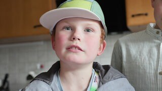 Ben, Spitzname "Lumpi", guckt in die Kamera. Er hat blaue Augen und eine gelb-grüne Kappe, unter der rötliche Haare hervorschauen. Im Hintergrund sieht man verschwommen eine Küche.