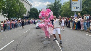 Eine Dragqueen in pinkem Kleid und pinker Kopfbedeckung läuft zwischen Zuschauern des CSD 2025 durch Stuttgart.