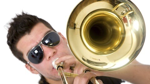 Junger Mann mit Sonnenbrille bläst in eine Posaune: Die Wucht von Trompete, Posaune und Tuba, moderne Rhythmen und Elemente von Techno bis Pop – neue Blasmusik-Combos sind mitreißend.