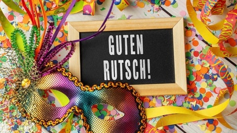Silvesterparty Ideen: Auf einem Tisch sieht man Konfetti und Luftschlangen und eine glitzernde Maske. Dazischen liegt eine kleine Tafel, auf der geschrieben steht: Guten Rutsch.