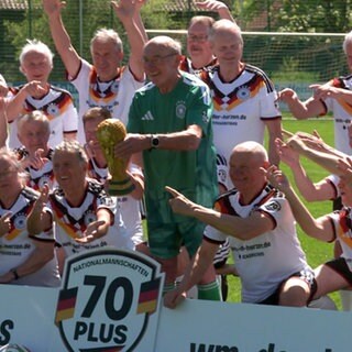 Die Spieler der Ü70-Fußball-Nationalmannschaft stehen hinter einer Bande und zeigen in die Mitte, wo ein älterer Mann in einem grünen Torwart-Outfit den WM-Pokal hält. Der Rest der Spieler trägt weiße Trikots mit schwarz-rot-goldenen Akzenten.