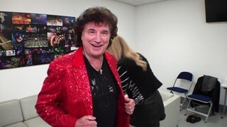 Olaf der Flipper trägt eine rote Anzugsjacke und lächelt in die Kamera. Im Hintergrund steht seine Tochter Pia. Sie stehen in einem weißen Raum in der Mannheimer SAP Arena.
