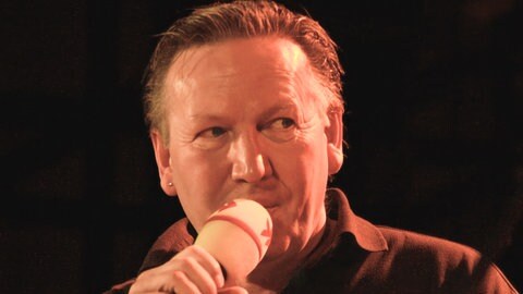 Matthias Holtmann mit Mikrofon auf der Bühne beim Finale der SWR1 Hitparade 2008.  Zum Tod von Matthias Holtmann: Wie erinnern sich Stars aus der Musikbranche, Weggefährten und SWR1 Hörer:innen an die Radiolegende und den Lebensbegleiter für viele Menschen?