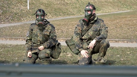 Eine junge Frau und ein junger Mann wollen Pflegesoldaten werden. Sie trainieren in Kampfanzug und mit Gasmaske.