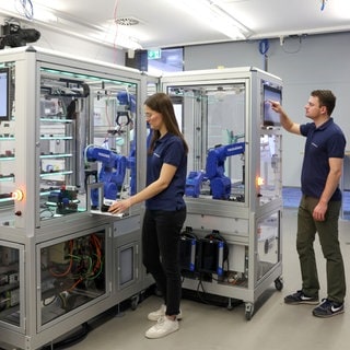 Eine Frau und ein Mann des Forschungsvereins Smart Factory stehen vor einer automatisierten Produktionsmaschine mit Greifarmen. Der Mann bedient ein Tablet, das Teil der Maschine ist.