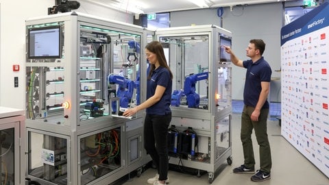 Eine Frau und ein Mann des Forschungsvereins Smart Factory stehen vor einer automatisierten Produktionsmaschine mit Greifarmen. Der Mann bedient ein Tablet, das Teil der Maschine ist.