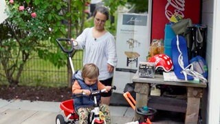 Nadine ist eine Mama mit Kleinwuchs. Gemeinsam mit einem ihrer Söhne ist sie draußen unterwegs und schiebt ihn auf einem Dreirad vor sich her.