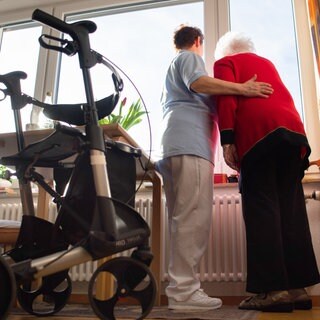 Eine Pflegerin und eine Seniorin stehen am Fenster, im Vordergrund ist ein Rollator zu sehen. Derzeit wird diskutiert, ob der Pflegegrad 1 abgeschafft werden soll, um Geld zu sparen.