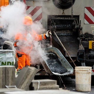 Dichter Dampf steigt auf, während ein Arbeiter heißen Asphalt aus der Maschine in die Schubkarre füllt. Auch die Bauwirtschaft leidet unter den Folgen des Irankriegs. (Symbolbild)