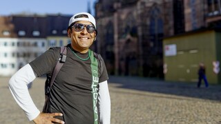 Samuel trägt eine Sonnenbrille und ein weißes Basecap. Er grinst frech in die Kamera und posiert mit einer Hand in der Hüfte aufgestützt.