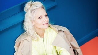 Ina Müller sitzt auf dem Boden in gelbem Oberteil und brauner Jacke über den Schultern