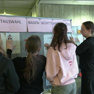 Vier Schülerinnen hängen Zettel an einer Pinnwand auf mit der Überschrift "Landtagswahl Baden-Württemberg 2026"