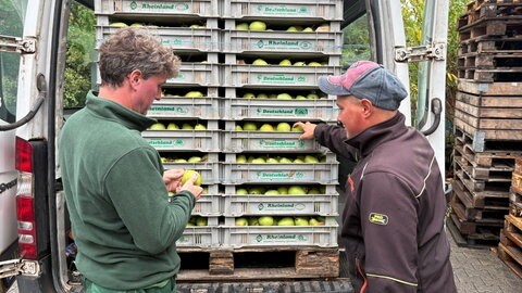 Obstbauer Gottschalk und ein Erntehelfer stehen vor einem mit Obstpaletten beladenen Transporter