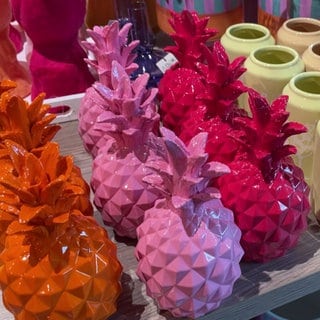 Mehrere orangene, pinke und lilane Ananas-Vasen stehen auf einem Tisch. Rechts daneben stehen schlichte Vasen in verschiedenen Farben.