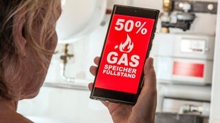 Rentnerin in ihrem Heizungsraum, auf dem Smartphone die alarmierende Nachricht: 50% Gasspeicher-Füllstand in Deutschland, sie fragt sich besorgt: Reicht das Gas im kommenden Winter? Steigen die Heizkosten?