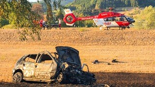 Ein ausgebranntes Fahrzeug im Vordergrund. Im Hintergrund ein Rettungshubschrauber. Durch den Unfall am Samstagabend geriet ein Fahrzeug in Brand. Das Feuer griff danach auf die Vegetation neben der Straße aus.