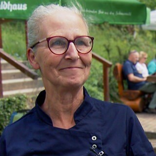 Silke ist Pächterin eines Wanderheims im Schwarzwald. Sie trägt eine Brille und eine dunkelblaue Kochjacke. Sie steht auf der Terrasse ihres Wanderheims. Im Hintergrund sieht man eine Gruppe Menschen an einem Holztisch sitzen.