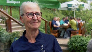 Silke ist Pächterin eines Wanderheims im Schwarzwald. Sie trägt eine Brille und eine dunkelblaue Kochjacke. Sie steht auf der Terrasse ihres Wanderheims. Im Hintergrund sieht man eine Gruppe Menschen an einem Holztisch sitzen.