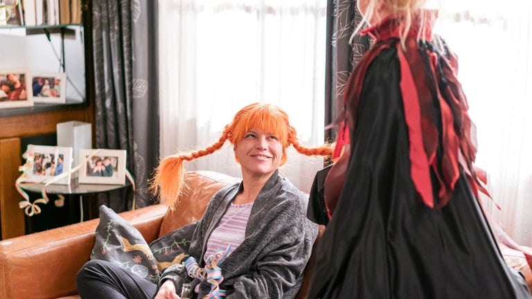 Eva (Lucie Muhr) sitzt auf dem Sofa, sie trägt eine Pipi-Langstrumpf-Perücke, Carlotta (Mila Breiter) steht vor ihr im Teufelchenkostüm