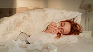Frau liegt mit Erkältung im Bett, putzt ihre Nase, tags: was Erkältungszeit Erkältung