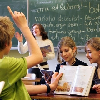Vor einer mit lateinischen Sprichwörtern beschriebenen Tafel erteilt eine Lehrerin Lateinunterricht. Schülerinnen und Schüler arbeiten in einer Gruppe: Das Schulfach Latein hat nicht den besten Ruf. Doch die alte Sprache bastelt an einer neuen Zukunft. Weniger elitär, mehr Geschichte und Alltag. Welches Potenzial steckt in Latein?