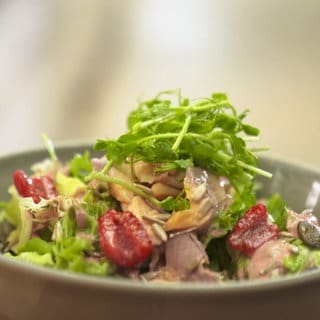Ein Salat mit Forellenstücken und getrockneten Tomaten liegt in einer hellen Schüssel.