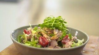 Ein Salat mit Forellenstücken und getrockneten Tomaten liegt in einer hellen Schüssel.