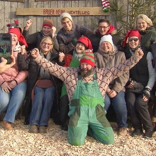 Bauer Heini steht mit seinem Team vor einem Holzgebäude. Alle tragen Weihnachtsmützen und freuen sich. Im Vordergrund macht eine Person mit einem Handy ein Foto.