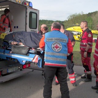 Eine Gruppe von Rettungskräften, darunter auch Personal der Heli-Retter, steht vor einem Krankentransport. Eine Trage wird eingeladen. Im Hintergrund steht der gelbe Helikopter "Christoph 22".
