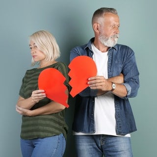Grauhaariges Paar, Mann und Frau, stehen nebeneinernder mit abgewandten Gesichtern und halten ein zerrissens rotes Papierherz: Immer häufiger trennen sich Paare nach langer Ehe. Warum wächst die Zahl der "grauen Scheidungen"?