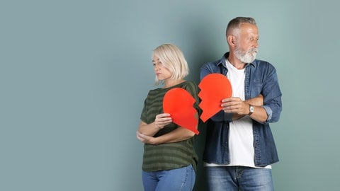 Grauhaariges Paar, Mann und Frau, stehen nebeneinernder mit abgewandten Gesichtern und halten ein zerrissens rotes Papierherz: Immer häufiger trennen sich Paare nach langer Ehe. Warum wächst die Zahl der "grauen Scheidungen"?